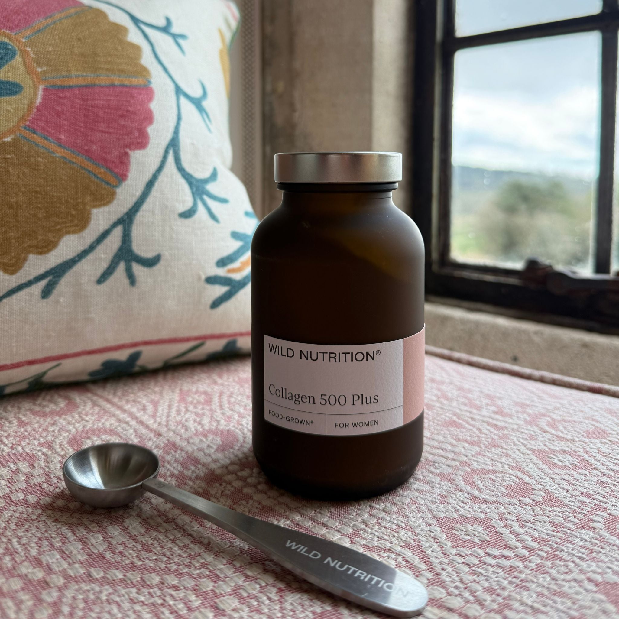 Wild Nutrition Collagen 500 Plus bottle with branded scoop on pink patterned cushion by a rustic window