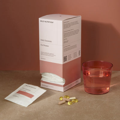 Wild Nutrition Daily Essentials for Women box with daily sachets, supplement capsules, and a glass of water on a warm-toned surface.